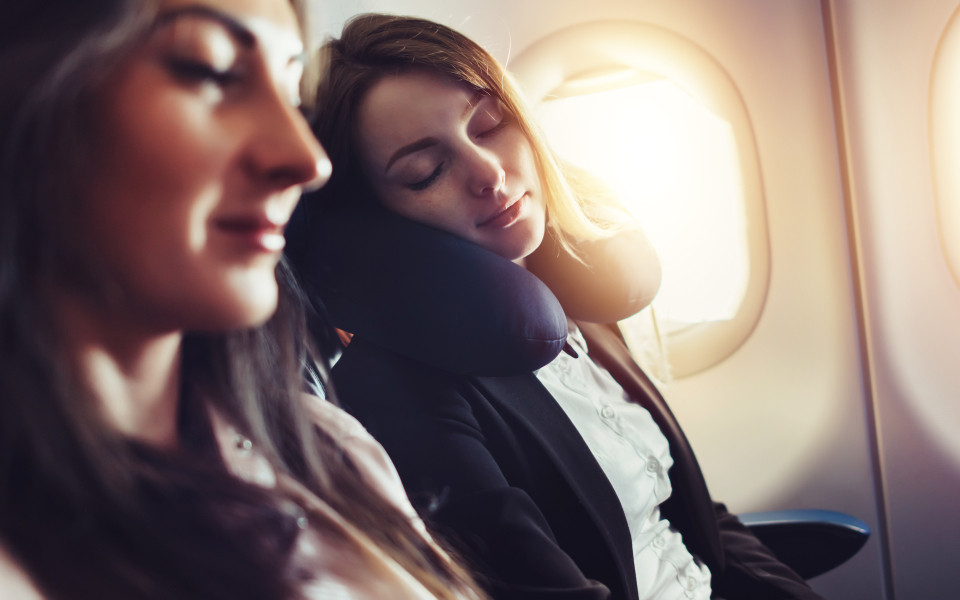 Eine Frau schläft entspannt mit einem Nackenkissen im Flugzeug, während die Sonne durch das Fenster scheint – ein Tipp zur Vermeidung von Jetlag.